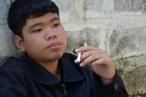 Boy learning to smoke by himself in the area behind the school fence which is a hidden place for people, bad influence of secondary school or junior high school life, addiction. photo