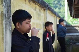 Boy learning to smoke by himself in the area behind the school fence which is a hidden place for people, bad influence of secondary school or junior high school life, addiction. photo