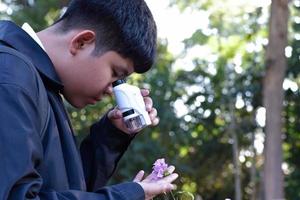 Young Asian boy is holding a pocket microscope to look at tiny particles on plant leaves to study plants and organisms on different types of plants in botanical garden, soft and selective focus. photo