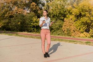 Happy young woman with coffee listening music on smartphone and walking on promenade photo