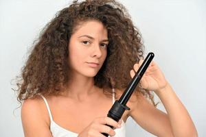 bella mujer sonriente con el pelo largo y ondulado planchándolo, usando un rizador. chica feliz con un hermoso cabello suave y saludable usando un rizador para rizos perfectos. peinado y peluqueria foto