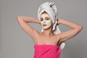 Beautiful female model with towel on her hear and white facial cosmetic mask on her face. Beauty cosmetic concept. Isolated on gray background. photo