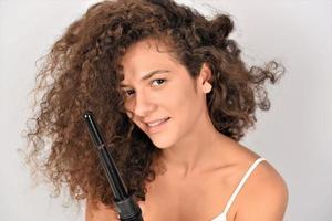 Pelo RIZADO. bella mujer sonriente con el pelo largo y ondulado planchándolo, usando un rizador. chica feliz con un hermoso cabello suave y saludable usando un rizador para rizos perfectos. peinado y peluqueria foto