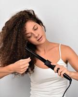 Beautiful Smiling Woman With Long  Wavy Hair Ironing It, Using Curling Iron. Happy Girl With Gorgeous Healthy Smooth Hair Using Curler For Perfect Curls. Hairstyle And Hairdressing photo