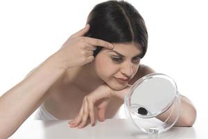 beautiful attractive brunette woman checking her face skin in front of a mirror on white background. photo