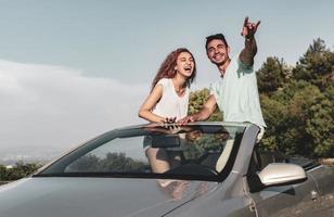 Friends having fun at car trip around the world. Couple in love with arms up on a convertible car. photo