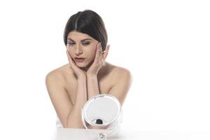 beautiful attractive brunette woman checking her face skin in front of a mirror on white background photo