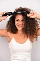 bella mujer sonriente con el pelo largo y ondulado planchándolo, usando un rizador. chica feliz con un hermoso cabello suave y saludable usando un rizador para rizos perfectos. peinado y peluqueria foto