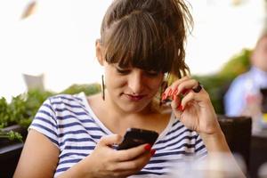 chica enviando mensajes de texto en el teléfono inteligente en la terraza de un restaurante con un fondo desenfocado