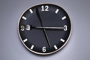 Clock on the black plate, with cutlery, grey background. The concept of intermittent fasting, lunchtime, diet and weight loss. photo