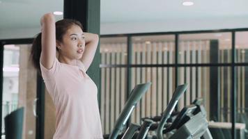 asiatische Frau, die sich die Haare schneidet, um sich auf ein Training im Fitnesscenter vorzubereiten. Frau trainiert auf Ellipsentrainer für Oberschenkelübungen. Fitness- und Ernährungskonzept.