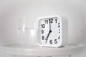 White alarm clock and glass of water on table in bedroom. Wake up early in the morning by the alarm bell and drink water. photo