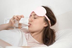 Young woman drinks a glass of water after waking up in the morning. Good habits for healthy way of life, dehydration concept, close up view. Diet, vitality, beauty care and healthy lifestyle. photo