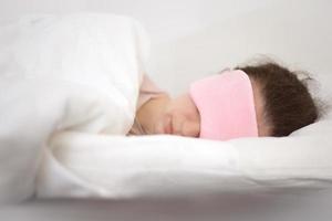 Young beautiful woman sleeping in bed with eye mask in the morning. Woman resting in comfortable white bed, lying on soft pillow, orthopedic mattress, enjoying good sleep. photo