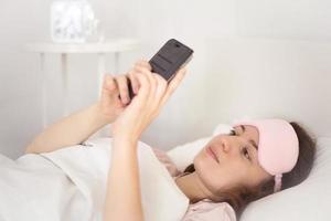 Relaxed young woman using a smartphone in the morning on the bed at home. Happy and smiling girl using a mobile phone. Woman texting, checking social apps, makes video call or playing with smartphone. photo