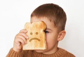 Children and gluten. Preschool boy with toasted bread, with sad emoji. Gluten intolerance by kids. photo