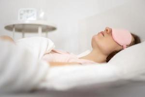 Young beautiful woman sleeping in bed with eye mask in the morning. Woman resting in comfortable white bed, lying on soft pillow, orthopedic mattress, enjoying good sleep. photo