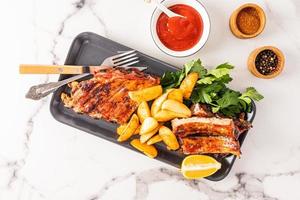 Top view of the ceramic dark tray with pork ribs grill and grilled potatoes with spices and herbs. marble white background. culinary menu. photo