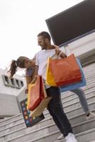 retrato de una alegre pareja joven caucásica hombre y mujer sosteniendo muchas bolsas de papel después de ir de compras mientras camina y habla en la calle. feliz pareja familiar con paquetes al aire libre. concepto de compra foto