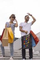 retrato de una alegre pareja joven caucásica hombre y mujer sosteniendo muchas bolsas de papel después de ir de compras mientras camina y habla en la calle. feliz pareja familiar con paquetes al aire libre. concepto de compra foto