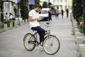 un hombre guapo está usando un teléfono inteligente y sonriendo mientras monta en bicicleta en la ciudad foto