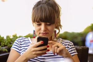 chica enviando mensajes de texto en el teléfono inteligente en la terraza de un restaurante con un fondo desenfocado