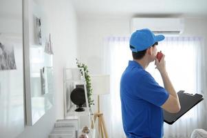 un joven técnico asiático de servicio que usa uniforme azul revisando, limpiando el aire acondicionado en casa foto