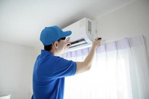 un joven técnico asiático de servicio que usa uniforme azul revisando, limpiando el aire acondicionado en casa foto