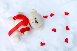 A toy bear with hearts lies on the ground against a snowdrift. photo