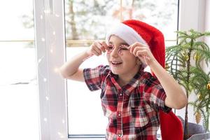 Girl in a Santa hat sits on the windowsill of a house near the Christmas tree and puts Candy cane to her eyes like glasses. Child is having fun and making faces, waiting for Christmas and New year photo