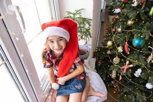 Happy girl in a Santa hat sits on the windowsill of a house near the Christmas tree. The child is having fun and making faces, waiting for Christmas and New year photo
