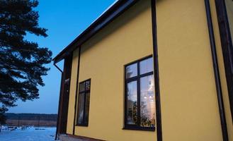 Windows of the yellow house are outside in the snow in winter. Inside the dwelling there is a warm light, fairy lights of garlands, a Christmas tree. Cozy celebration of Christmas and New Year at home photo