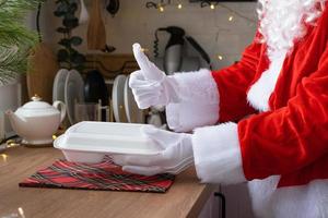 Food delivery to home service containers in hands of Santa Claus puts it on kitchen table and takes it away, thumbs up. Ready-made hot order, Christmas, New year holidays catering. photo