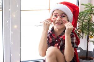 Girl in a Santa hat sits on the windowsill of a house near the Christmas tree and puts Candy cane to her eyes like glasses. Child is having fun and making faces, waiting for Christmas and New year photo