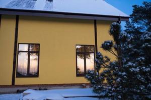 las ventanas de la casa amarilla están afuera en la nieve en invierno. dentro de la vivienda hay una luz cálida, luces de hadas de guirnaldas, un árbol de navidad. acogedora celebración de navidad y año nuevo en casa foto