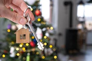 Key to the house in hand on background of Christmas tree. Gift for New Year, Christmas. Building, design, project, moving to new house, mortgage, rent and purchase real estate. Copy space