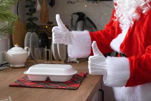 Food delivery to home service containers in hands of Santa Claus puts it on kitchen table and takes it away, thumbs up. Ready-made hot order, Christmas, New year holidays catering. photo