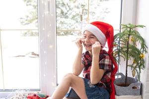 Girl in a Santa hat sits on the windowsill of a house near the Christmas tree and puts Candy cane to her eyes like glasses. Child is having fun and making faces, waiting for Christmas and New year photo