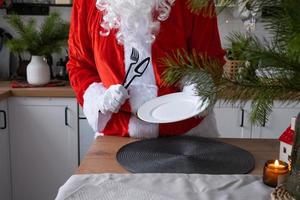 Santa Claus sets the table at home in the white kitchen, puts a plate, puts cutlery fork and knife. Preparing for the celebration of Christmas, setting the table for guests photo