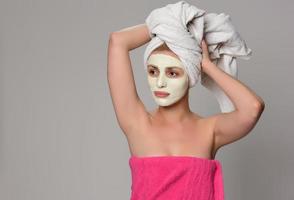 Beautiful female model with towel on her hear and white facial cosmetic mask on her face. Beauty cosmetic concept. Isolated on gray background. photo