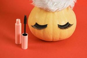 mascara in pink packaging mock up and pumpkin in santa hat and false eyelashes on red background photo