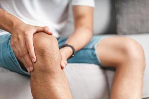 el hombre que sufre de dolor de rodilla sentado en el sofá sosteniendo y masajeando su rodilla dolorosa. foto