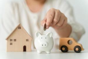 cierre de la mano de una mujer poniendo monedas en una alcancía blanca para ahorrar dinero, una casa de juguetes y un coche en la mesa, ahorrando dinero y concepto financiero foto