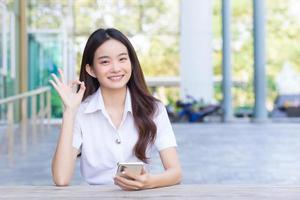 Young Asian woman students is sitting using smartphone to search information for a study report and signal an okay hand in university.