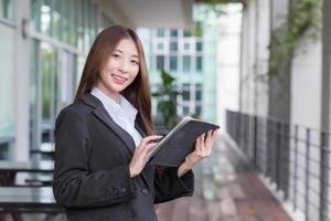 Asian professional business young woman in dark blue suit smiles happily stand with confidence and look at the camera while she works and holds tablet front office. photo