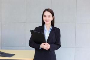 Young Asian professional working woman in a black suit holds clipboard in her hands and confident smiles in office room. photo