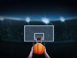 mano sosteniendo una pelota de baloncesto a punto de dispararla en el tablero de baloncesto hay un anfiteatro en el fondo. foto