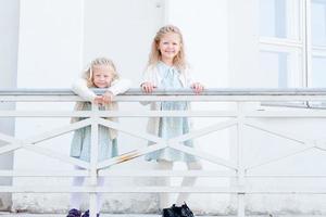 2 little blonde girls with curly hair are leaning on the railing on the balcony. Cute and little girls on the street balcony. 2 sisters walking outside in matching dresses and white tights photo