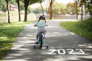 feliz año nuevo 2023,2023 simboliza el comienzo del nuevo año. la carta comienza el nuevo año 2023 en la bicicleta de ciclismo de la niña en la carretera en el jardín del parque natural. meta del éxito. fondo de pantalla del número 2023. foto