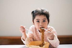 la niña asiática disfruta feliz usando cubiertos, cuchara y tenedor comiendo deliciosos fideos en la cocina en la mesa del comedor. una niña asiática feliz practica comer sola en la mesa del comedor. concepto de comida para bebés foto